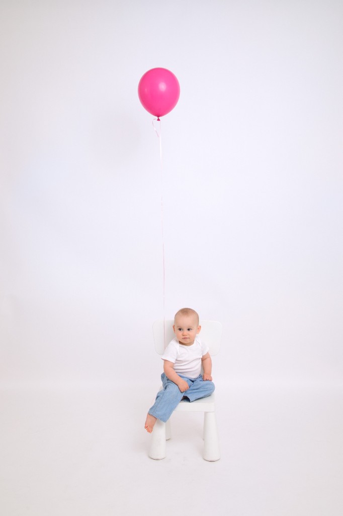 Milania, almost one year old, sitting on a small white chair with a pink balloon floating above her